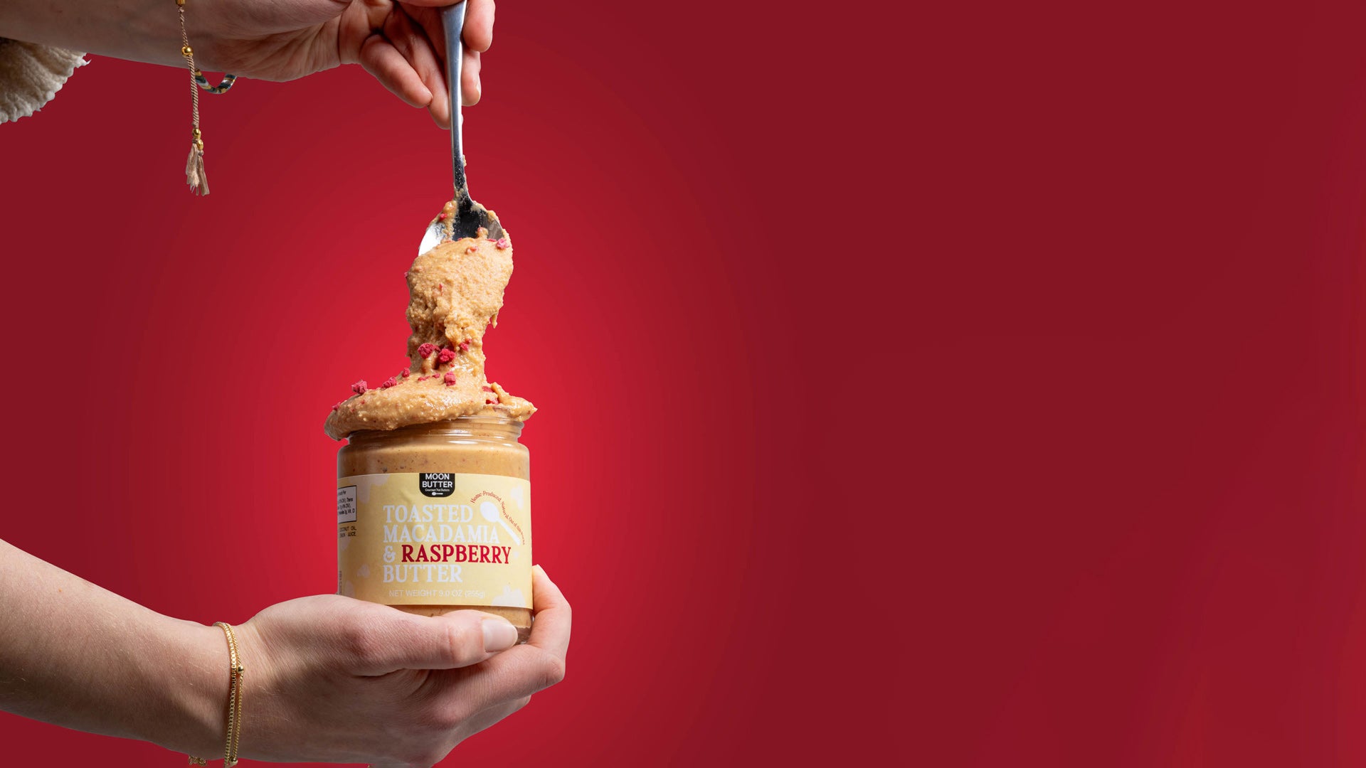 Person holding a jar of macadamia raspberry nut butter with a spoonful being lifted against a red background