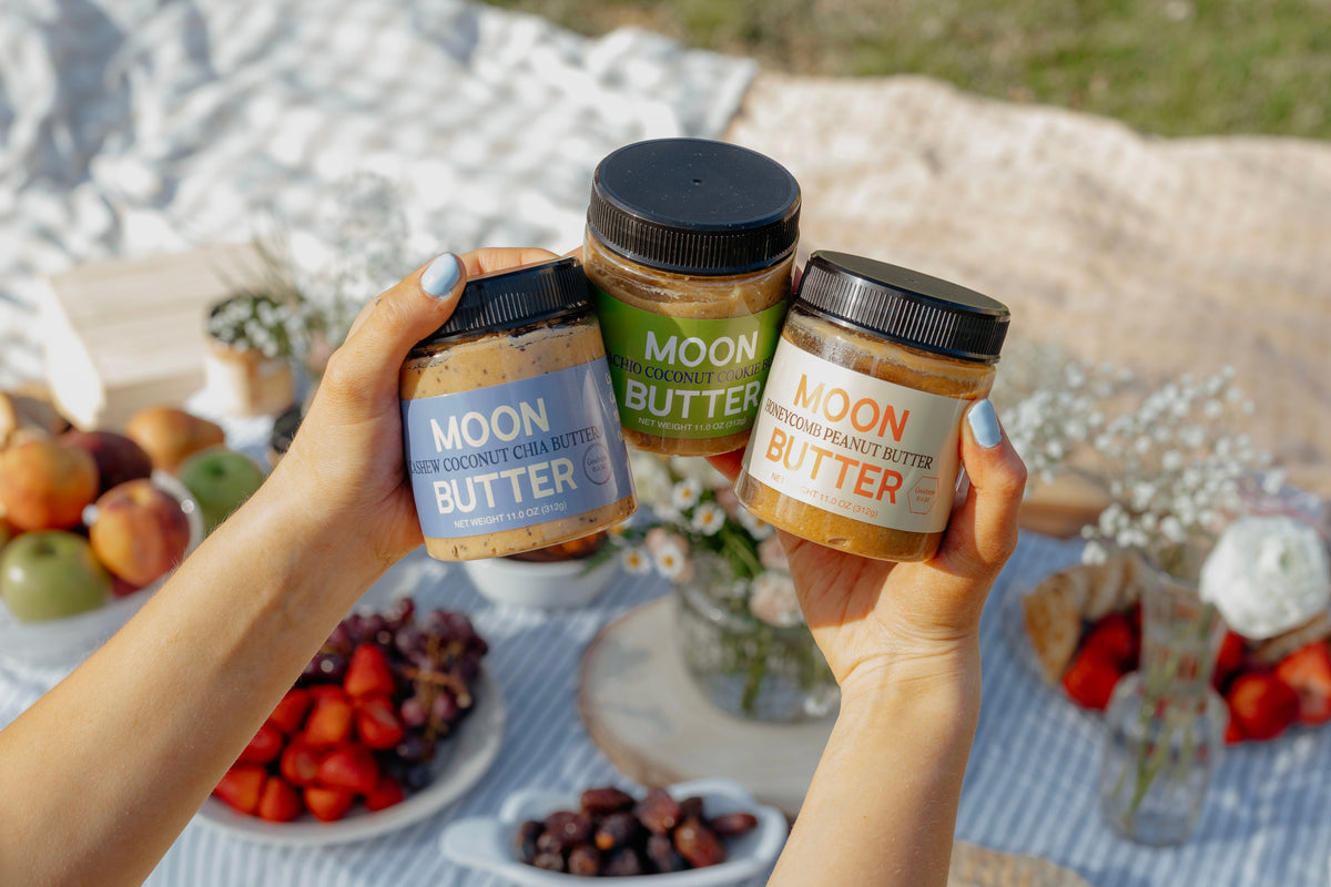 Hands holding three jars of Moon Butter: Cashew Coconut Chia, Pistachio Coconut Cookie, and Honeycomb Peanut Butter
