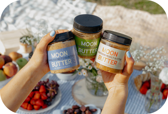 Hands holding three jars of Moon Butter: Cashew Coconut Chia, Pistachio Coconut Cookie, and Honeycomb Peanut Butter