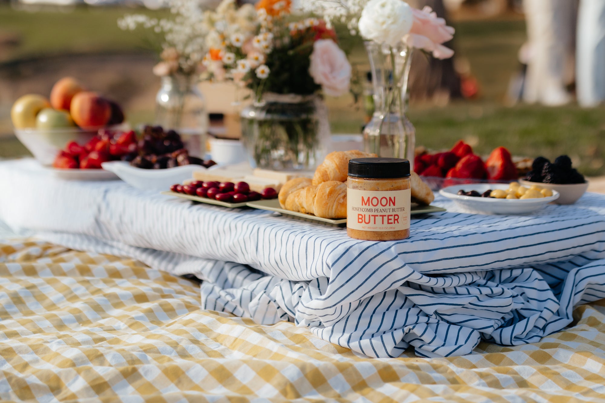 Moon Butter Honeycomb Peanut Butter on a striped picnic tablecloth with croissants, fruit, and flowers